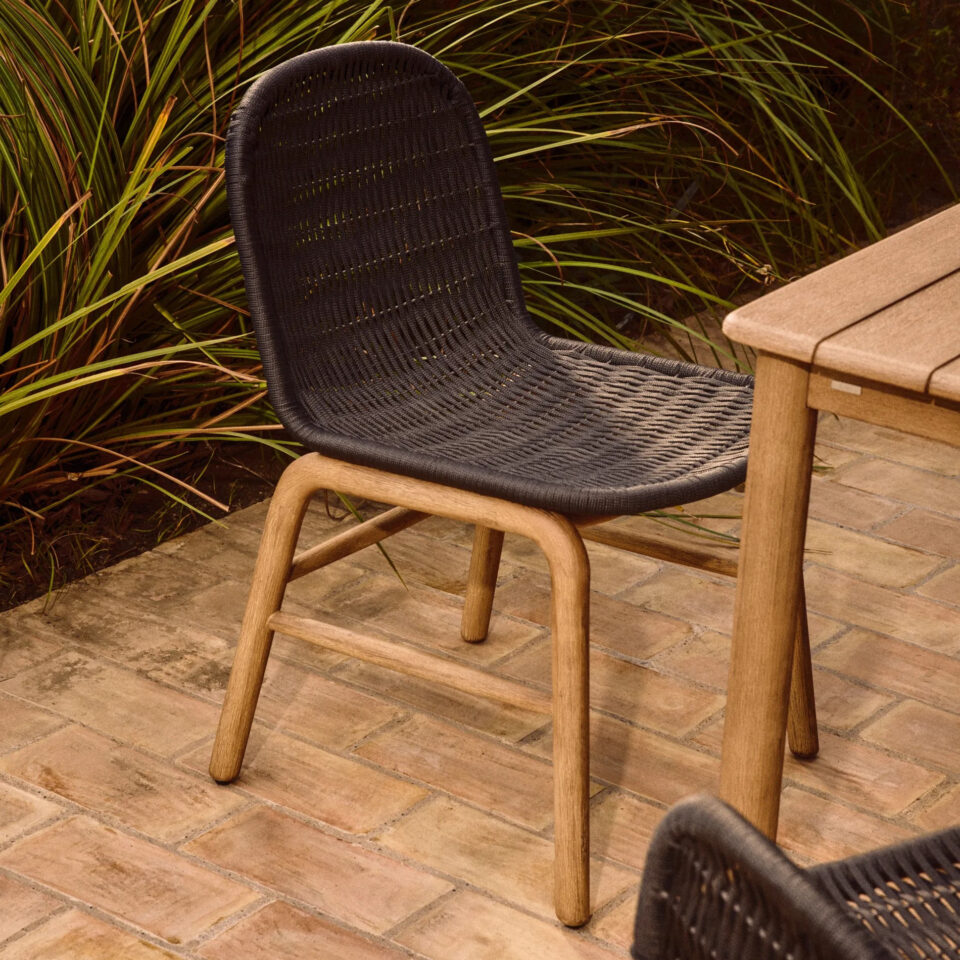 Črn vrtni stol Alipa v naravnem zunanjem okolju na terasi z opečnatimi ploščicami in zelenjem v ozadju, poleg lesene mize.