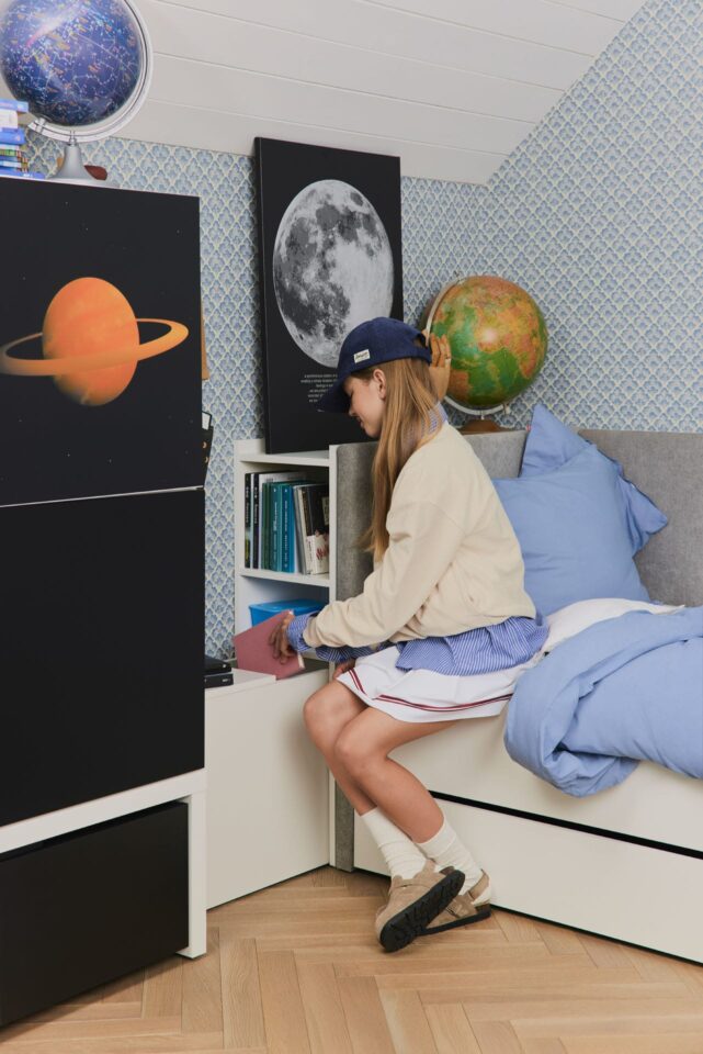 Deklica sedi na postelji ob regalu z nočno omarico